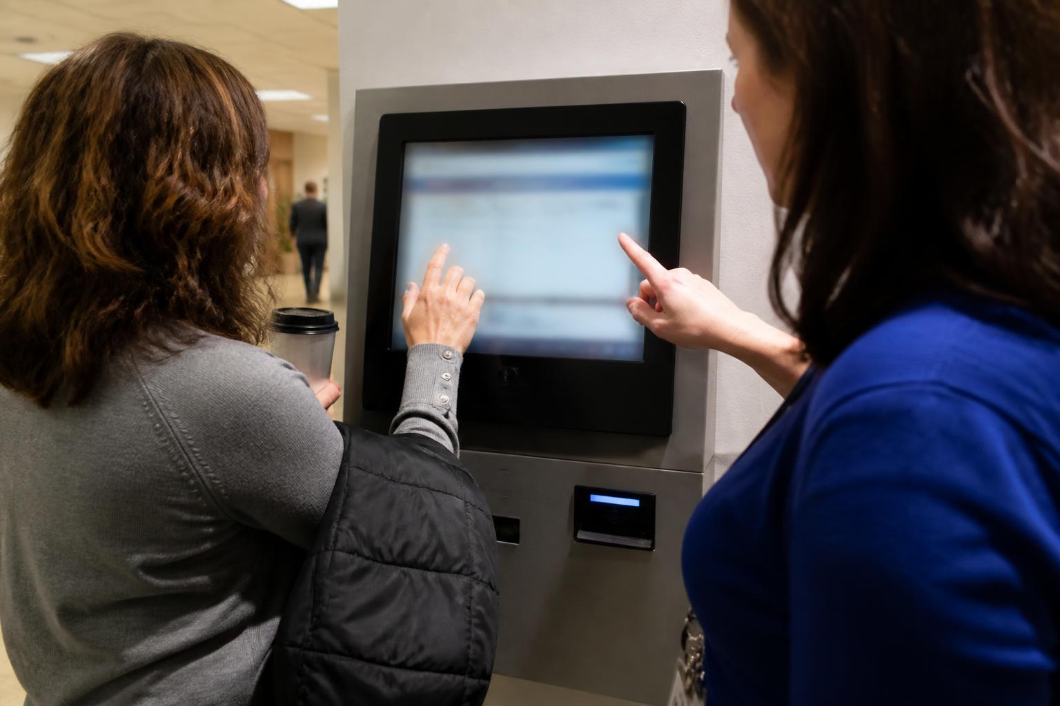 A worker adjusting a withholding setting at the kiosk while a payroll staffer points from the side.