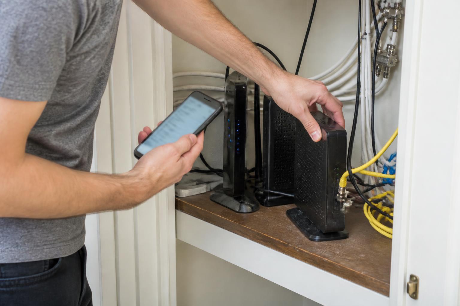 A renter unplugging an old modem while checking a blurred plan setting on a phone.