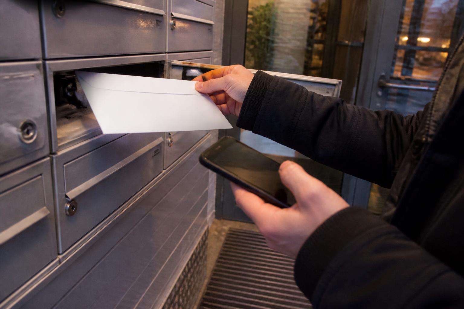 A borrower placing a sealed card-payment envelope into outgoing mail while checking a dark phone screen.