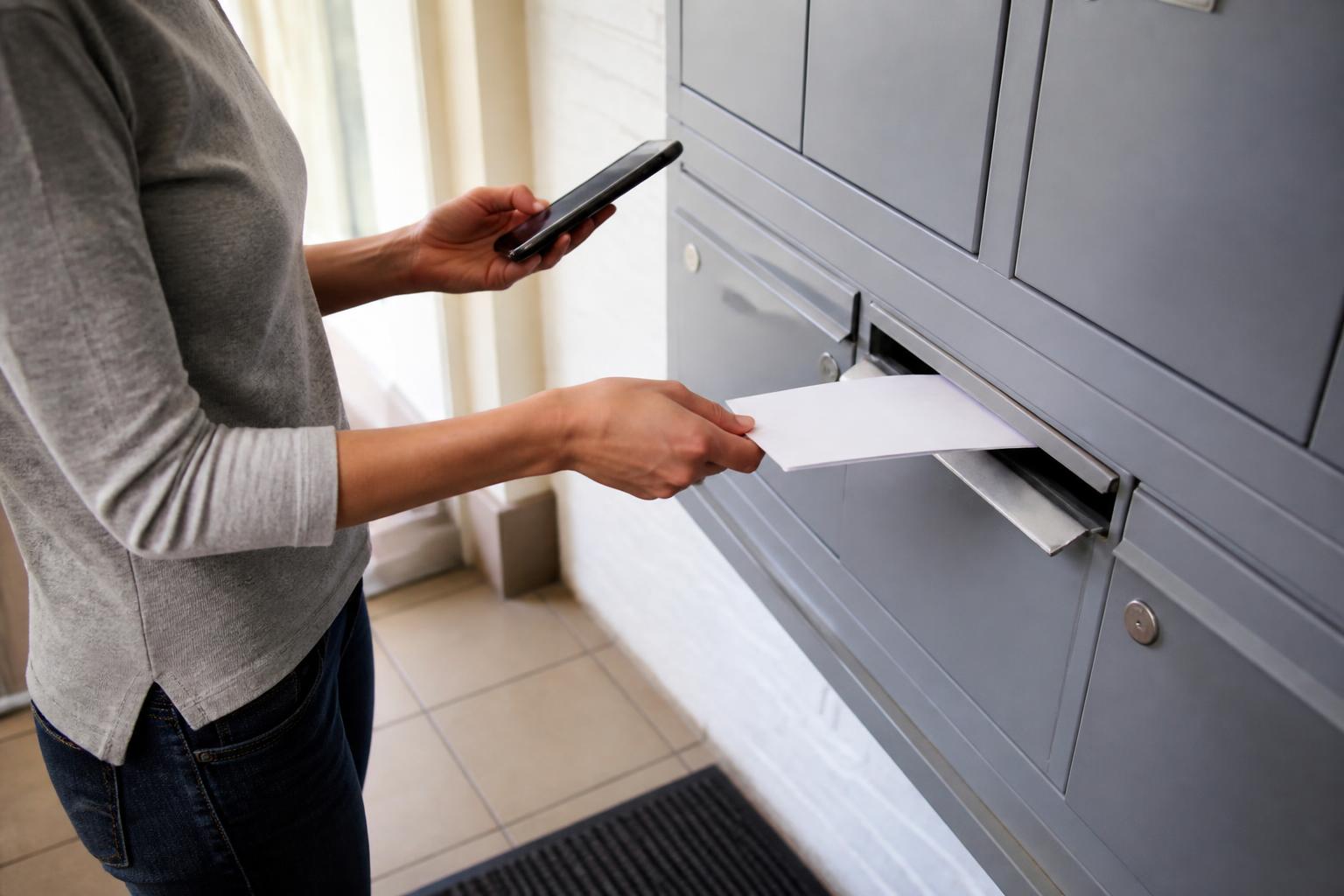 A borrower placing a sealed card-payment envelope into outgoing mail while checking a dark phone screen.