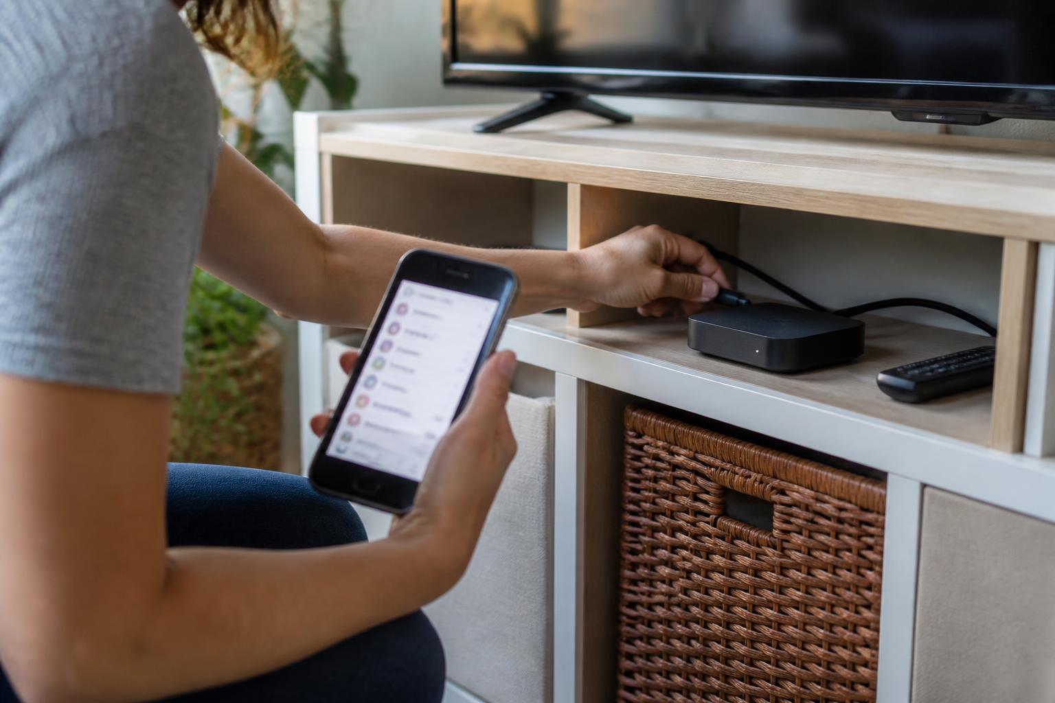 A person unplugging an unused streaming device while holding a phone with a blurred subscription settings screen.
