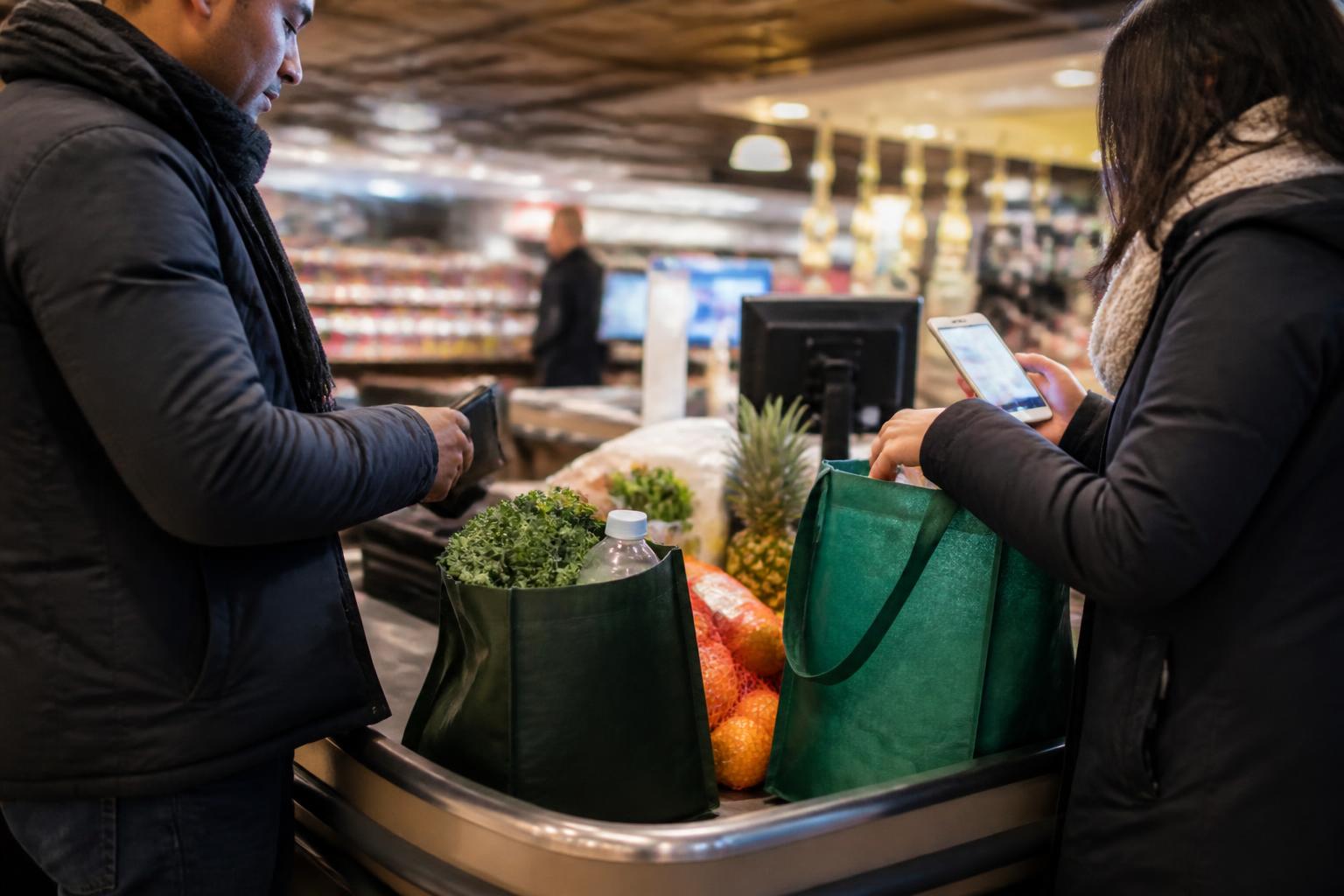 Two adults dividing groceries at checkout while checking a shared-spending alert.
